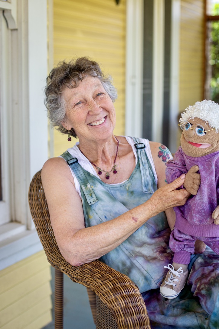 Carrie Elderberry seated on her porch with a grandmother puppet